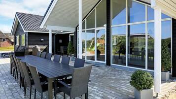 A modern outdoor dining area with a long table and chairs, large windows, and a brick patio.