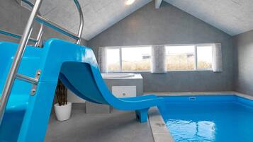 An indoor swimming pool with a blue slide, a window with curtains, and a view of the outdoors.