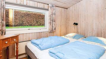 Un dormitorio con techo de madera, una ventana con vista a un seto, una mesita de noche y dos camas con ropa de cama azul.