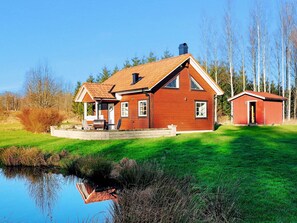 A two-story wooden house with a covered porch, a small shed, and a pond in the foreground. - 6 Person Holiday Home in Vittsjo (Vittsjo)