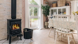 A cozy room with a wood-burning stove, a dining table with chairs, and a window overlooking a garden. - Holiday Home in Jerup (Jerup)