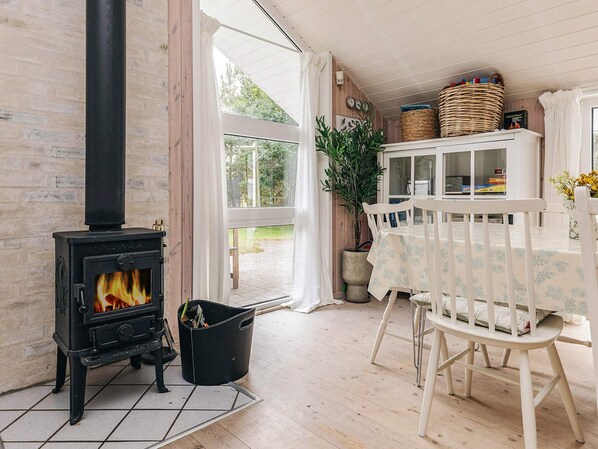 A cozy room with a wood-burning stove, a dining table with chairs, and a window with curtains.