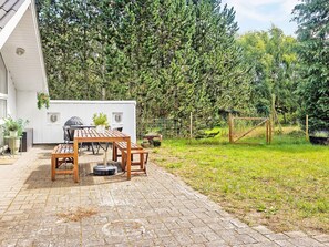 A patio area with a wooden table and chairs, a grill, and a white building with windows. - Holiday Home in Jerup (Jerup)