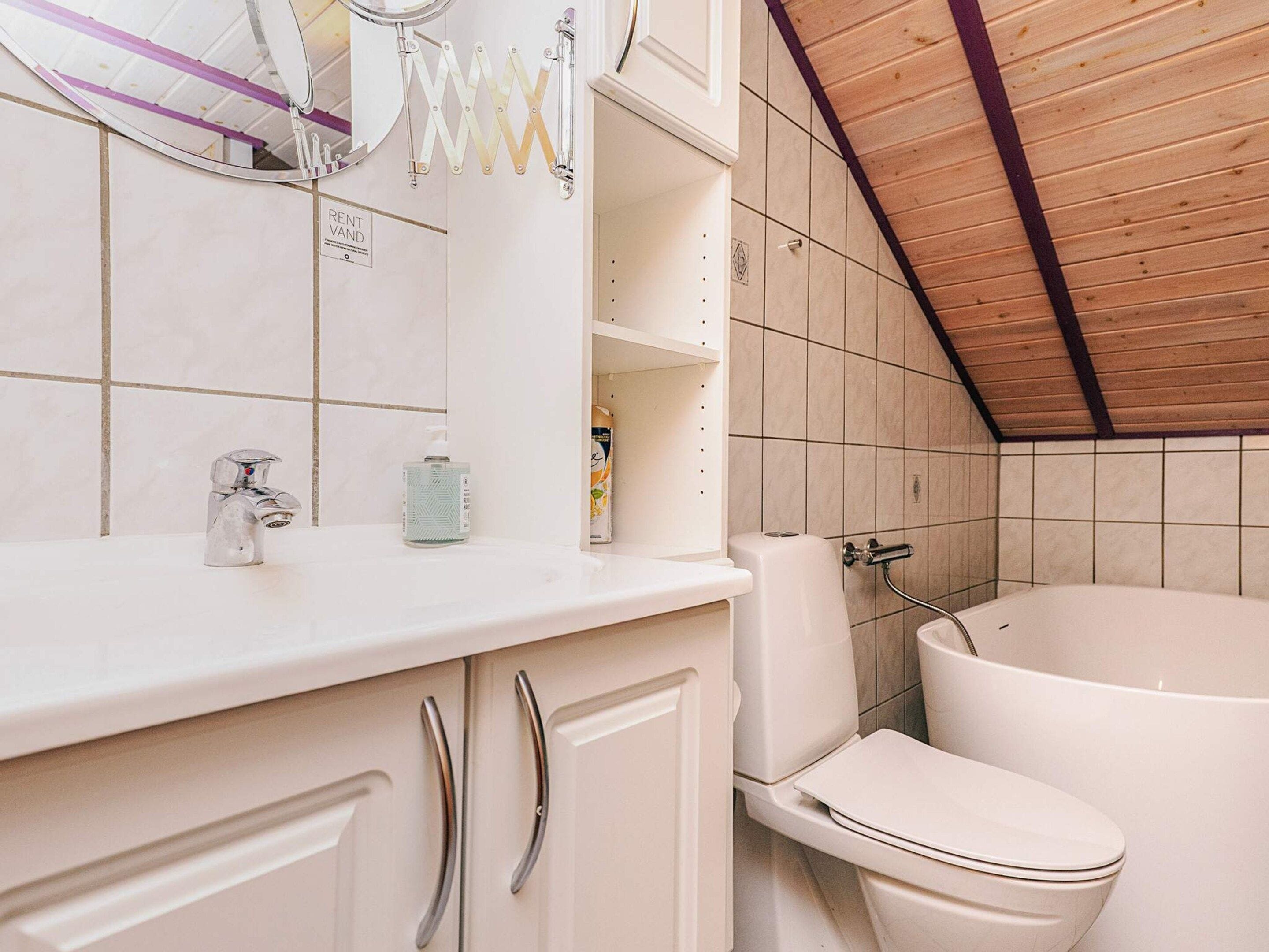 A bathroom with a white sink, mirror, and toilet. There is a wooden ceiling and tiled walls.