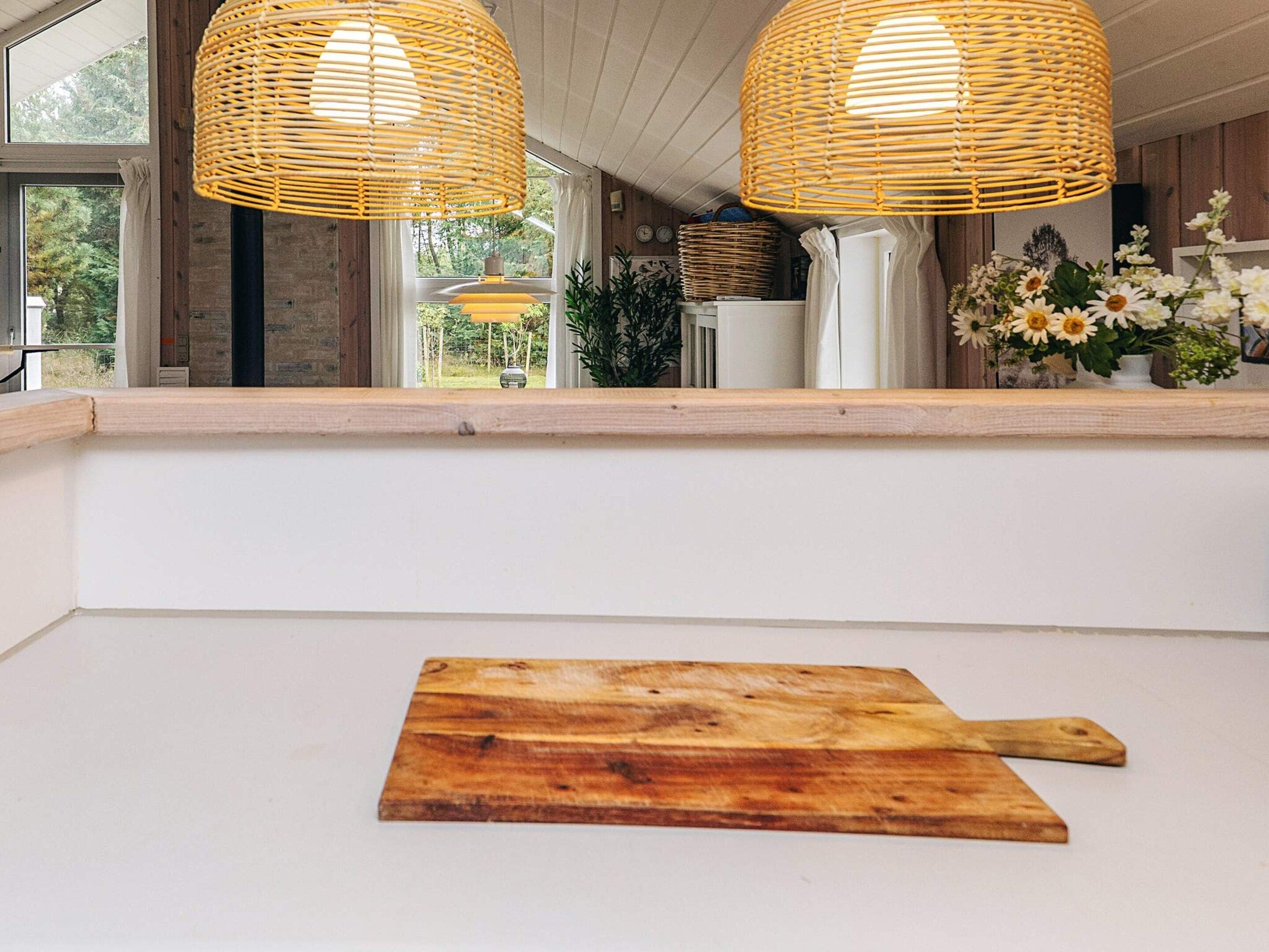 A kitchen with two wicker pendant lights, a wooden cutting board, and a window overlooking greenery.