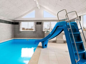 Une piscine intĂ©rieure avec un toboggan bleu, des murs recouverts de carreaux gris et de grandes fenĂȘtres.