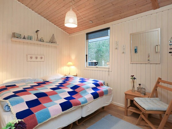 A bedroom with a bed, a window, a bedside lamp, a wooden chair, and a shelf with decorative items.