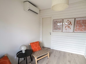 Quarto com parede branca, porta de madeira, cadeira de madeira pequena com almofada laranja, mesa redonda com abajur e aparelho de ar condicionado.