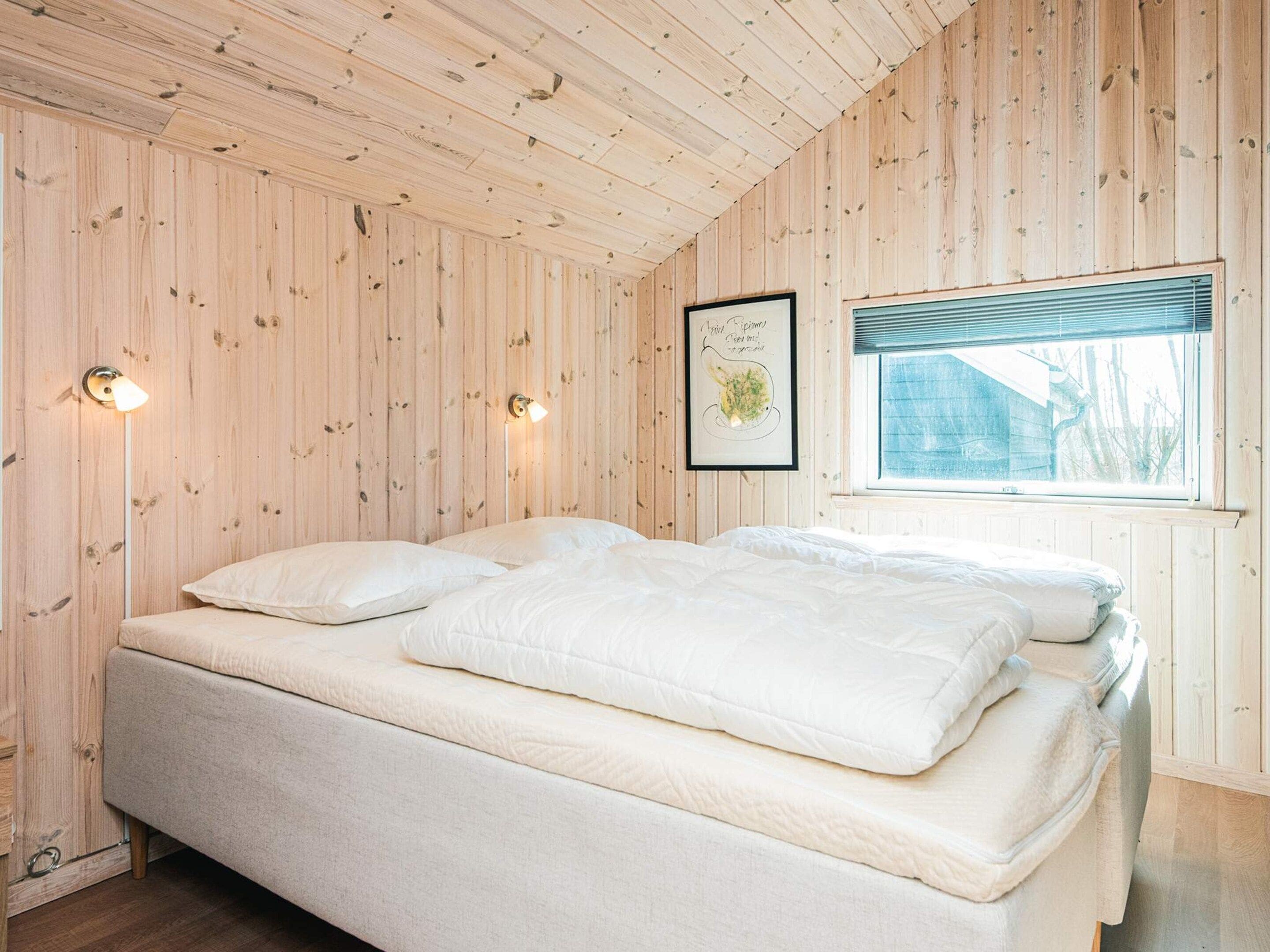A bedroom with a wooden paneled ceiling and walls, a bed with white bedding, two wall-mounted lamps, and a window with blinds.