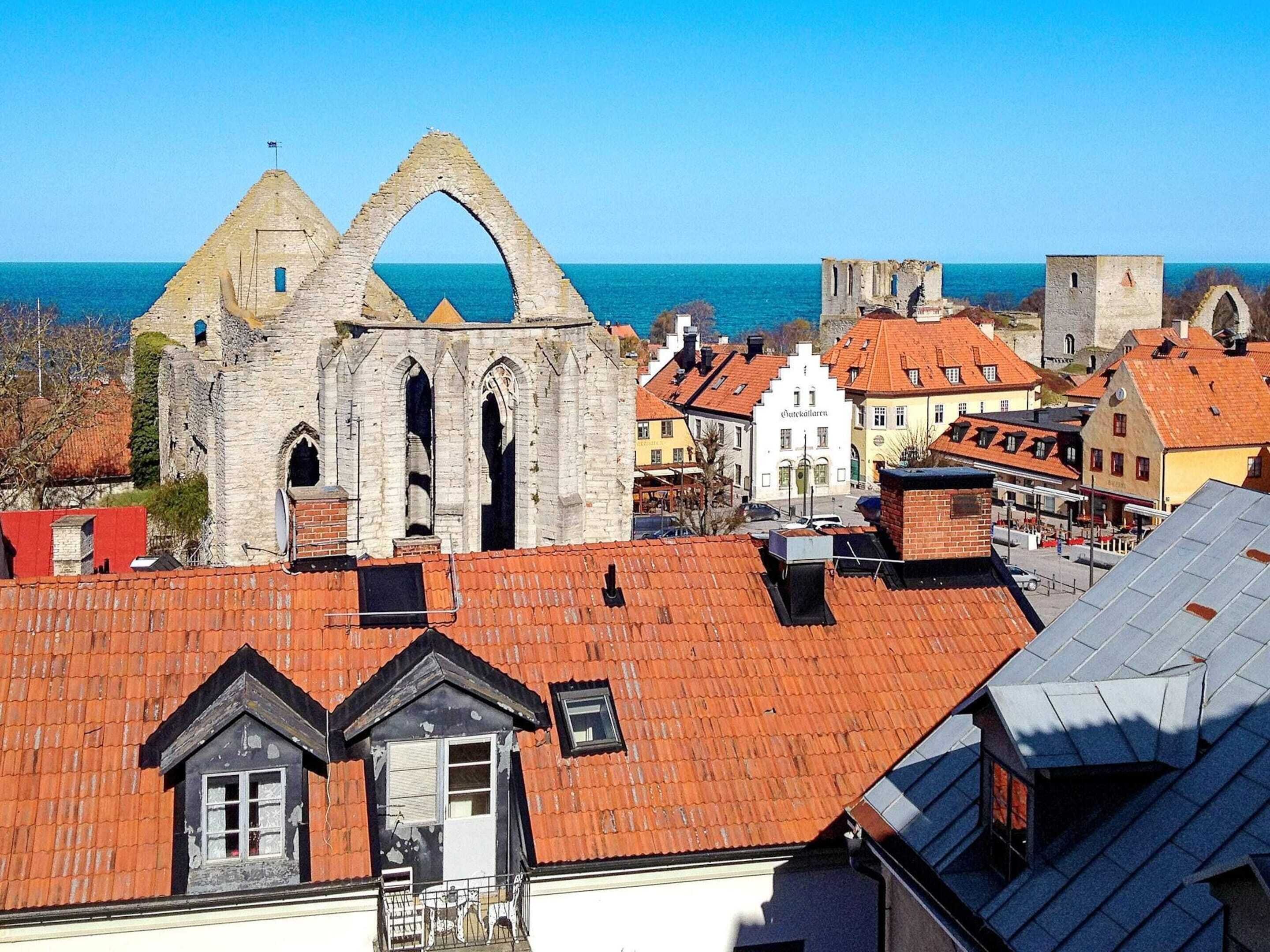 A historic stone building with a large archway, surrounded by traditional European architecture and a view of the sea.