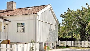 Une maison blanche à deux étages, avec un toit recouvert de tuiles rouges, un porche à l’avant et une clôture blanche faite de piquets.