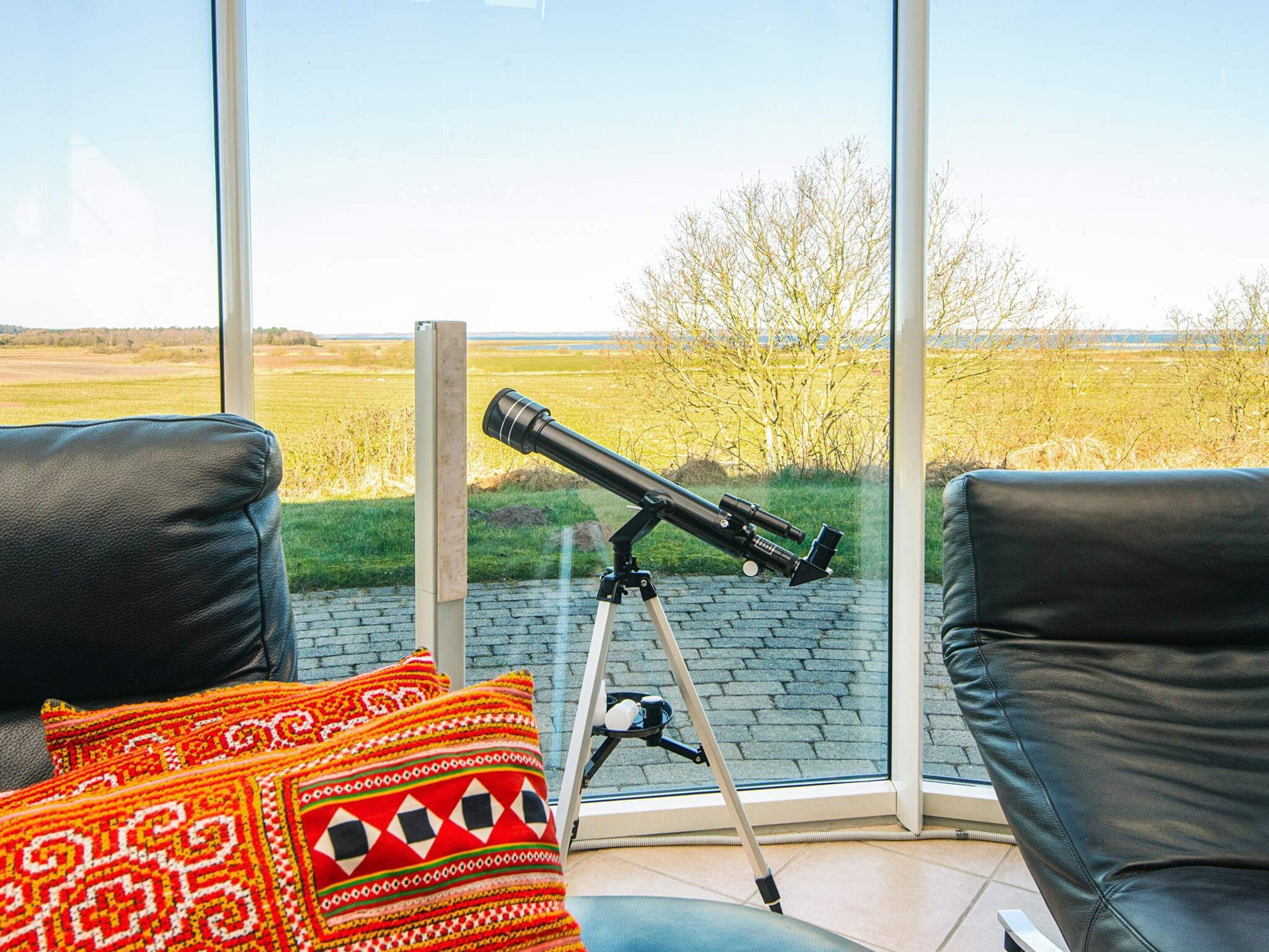 Un telescopio sobre un trípode en una habitación con ventanas amplias, sillones de cuero y cojines estampados.