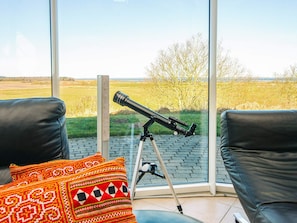 A telescope on a tripod in a room with large windows, leather chairs, and patterned cushions. - 5 Star Holiday Home in Hojslev (Hojslev)