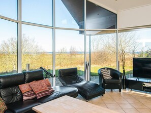 A modern living room with a large glass wall, leather furniture, and a flat-screen TV. - 5 Star Holiday Home in Hojslev (Hojslev)