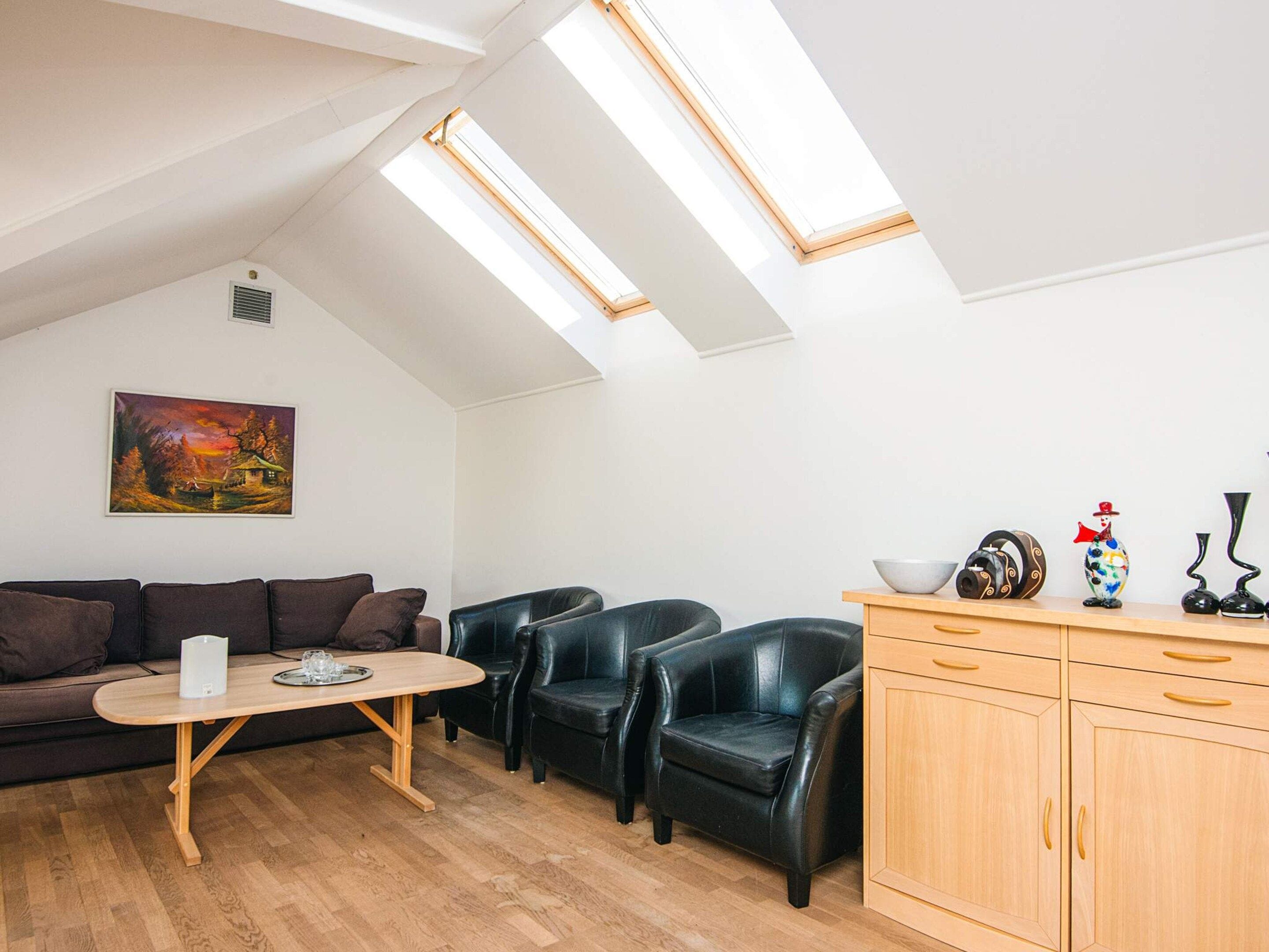 Una habitación estilo loft con techo inclinado, una mesa de comedor de madera, un sofá, dos sillones de cuero negro, un armario y un cuadro en la pared.