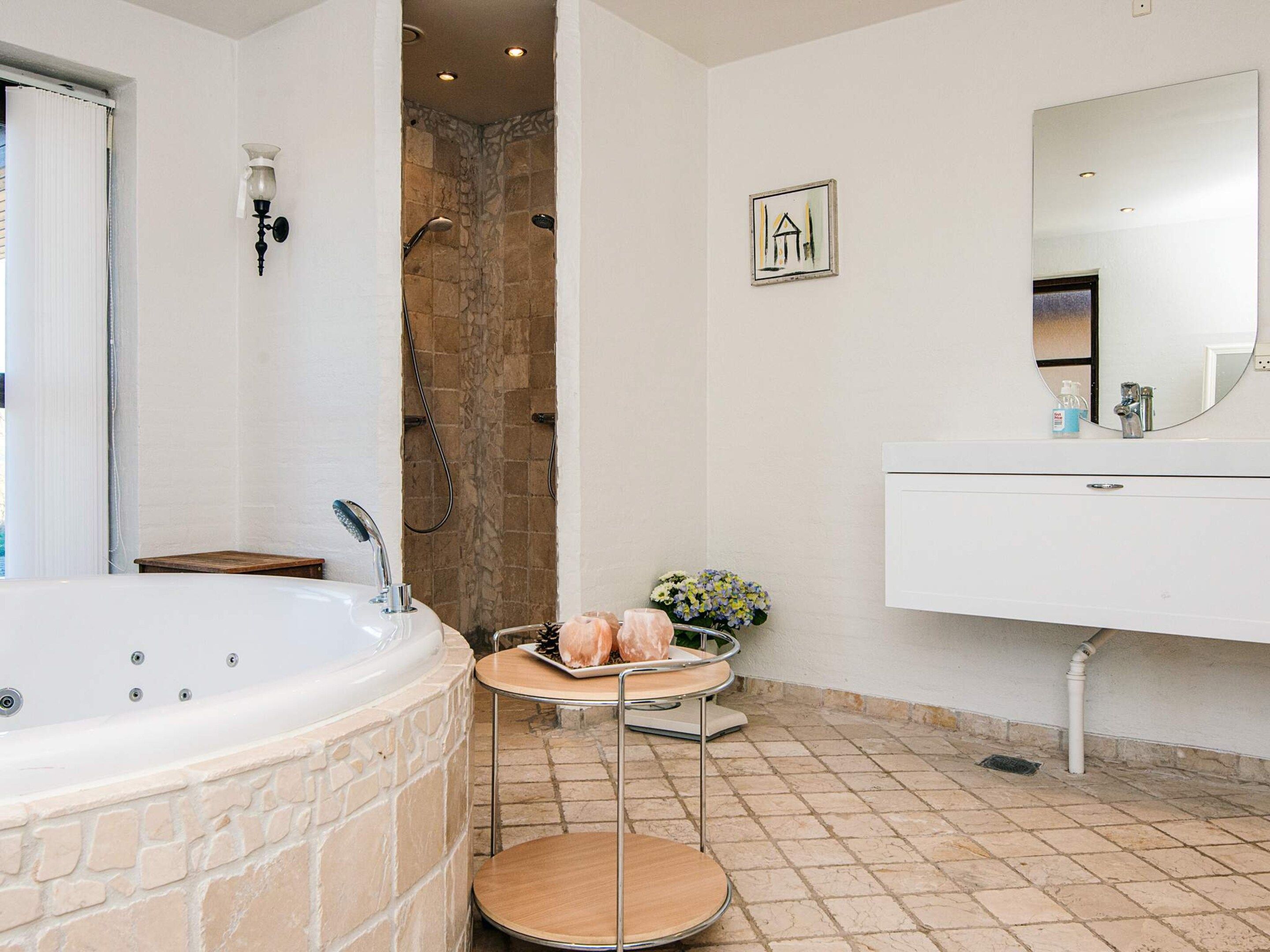 Une salle de bain avec une douche en pierre, un lavabo blanc avec un miroir et une petite table en bois sur laquelle sont posés des articles de toilette.