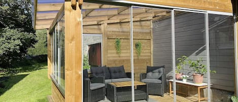 Una terraza de madera con mobiliario exterior, cerramiento de vidrio y vistas a un jardín.