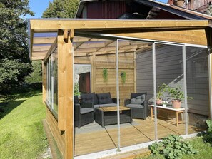 Una terraza de madera con mobiliario exterior, cerramiento de vidrio y vistas a un jardín.