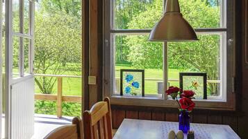 A dining area with a table, chairs, a hanging lamp, and a view of a green landscape through large windows.