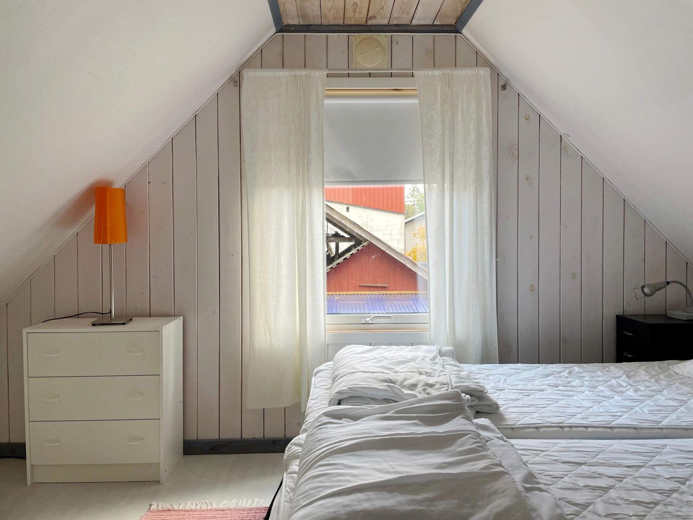 Un dormitorio con techo inclinado, cama, mesita de noche, lámpara y ventana con cortinas.