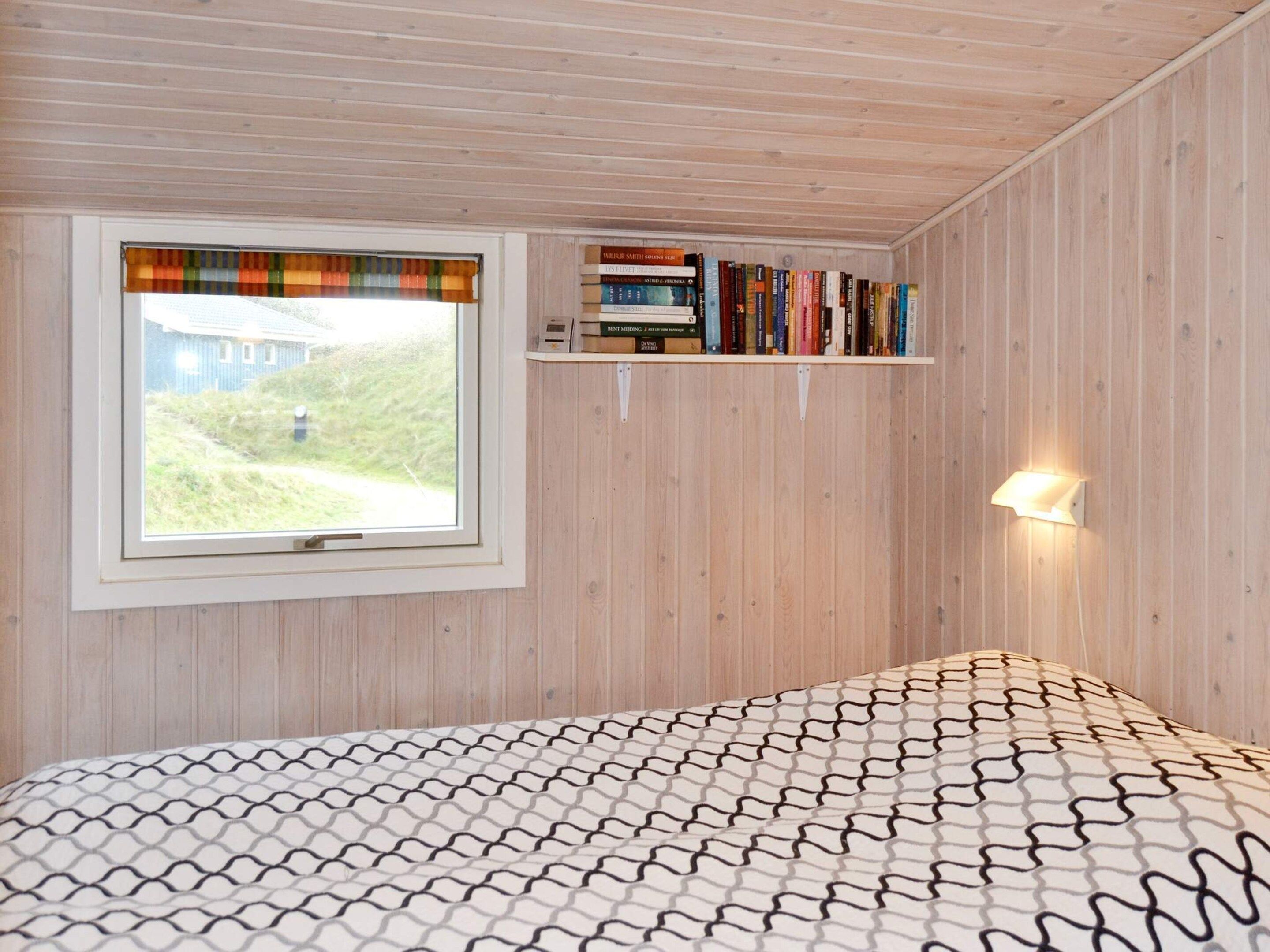 Una habitación con cama, ventana con vista a un edificio, estante con libros y una lámpara fijada a la pared.