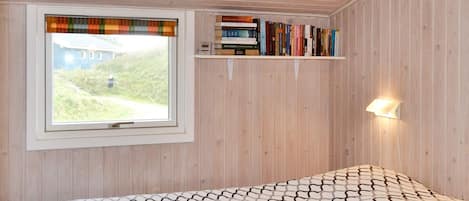 Una habitación con cama, ventana con vista a un edificio, estante con libros y una lámpara fijada a la pared.