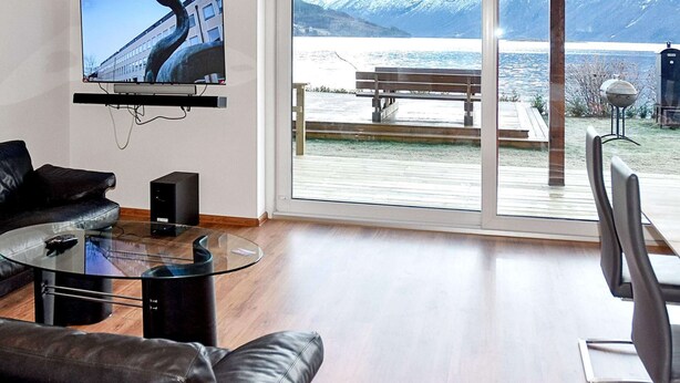 A modern living room with a large TV, leather furniture, and a view of a snowy mountain and lake. - 6 Person Holiday Home in Eidsvag (Molde)