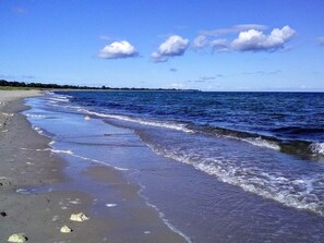 A beach with waves gently lapping onto the shore, a clear sky, and a distant landmass. - 5 Star Holiday Home in Vaeggerlose (Vaeggerlose)