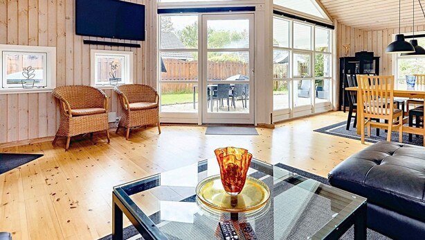 A spacious living room with wooden floors, a glass coffee table, wicker chairs, and a large sliding glass door leading to an outdoor patio. - 5 Star Holiday Home in Vaeggerlose (Vaeggerlose)