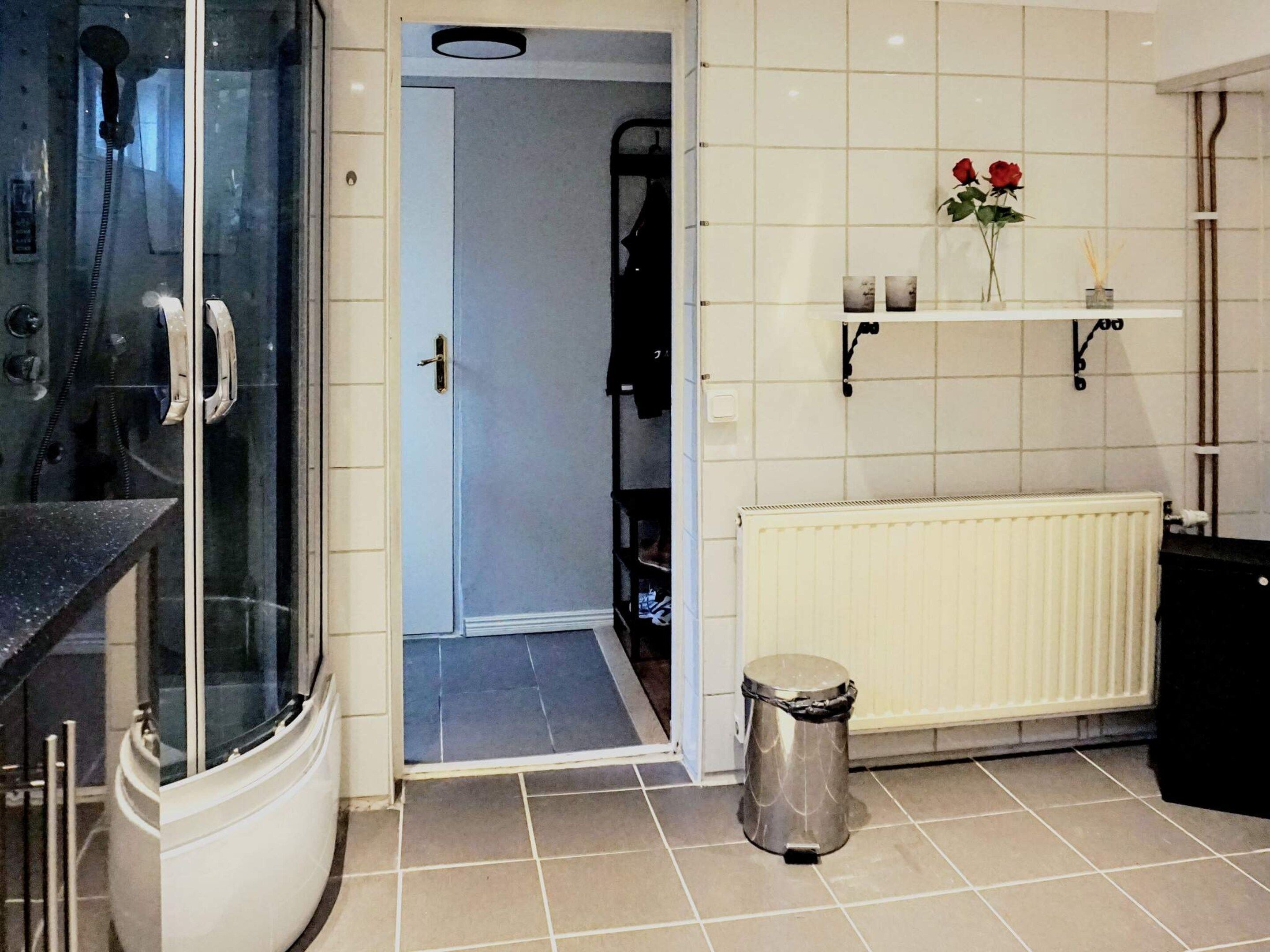 A bathroom with a shower, a radiator, a trash bin, and a shelf with flowers.