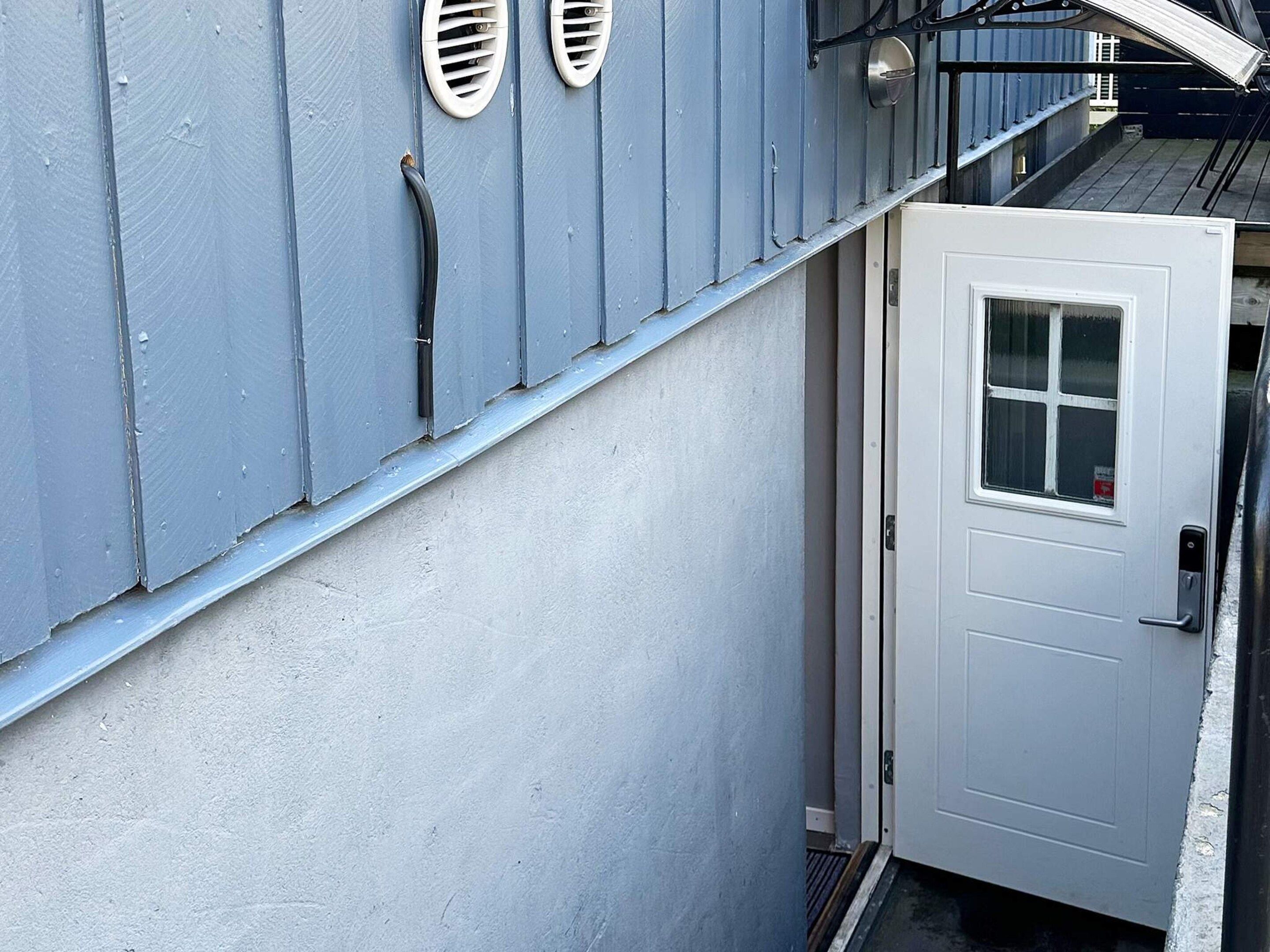 Une porte blanche avec une fenêtre, une rambarde en métal et un mur bleu percé de bouches d’aération.