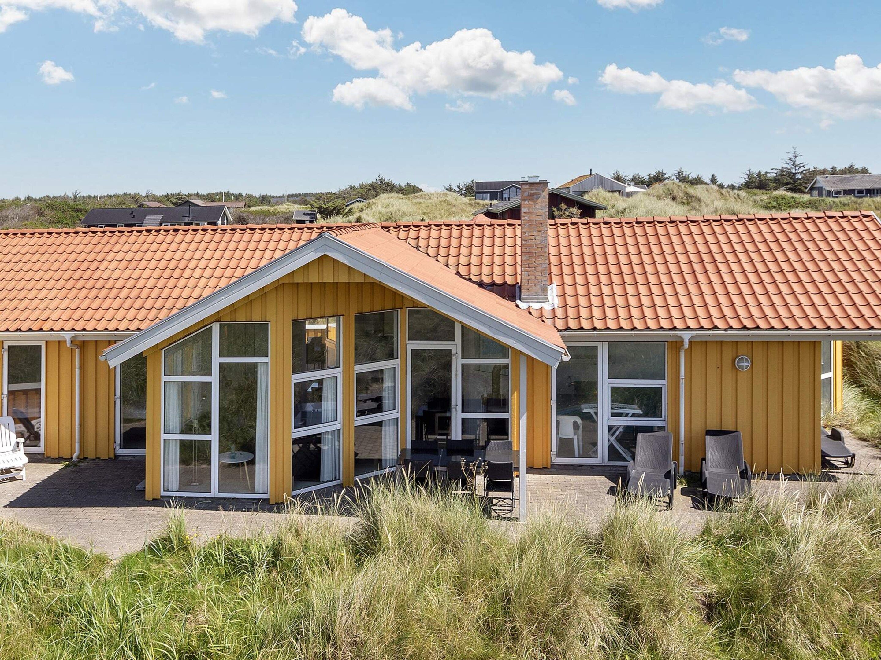 En gul bygning med rødt tegltag, store vinduer og udendørs siddepladser.