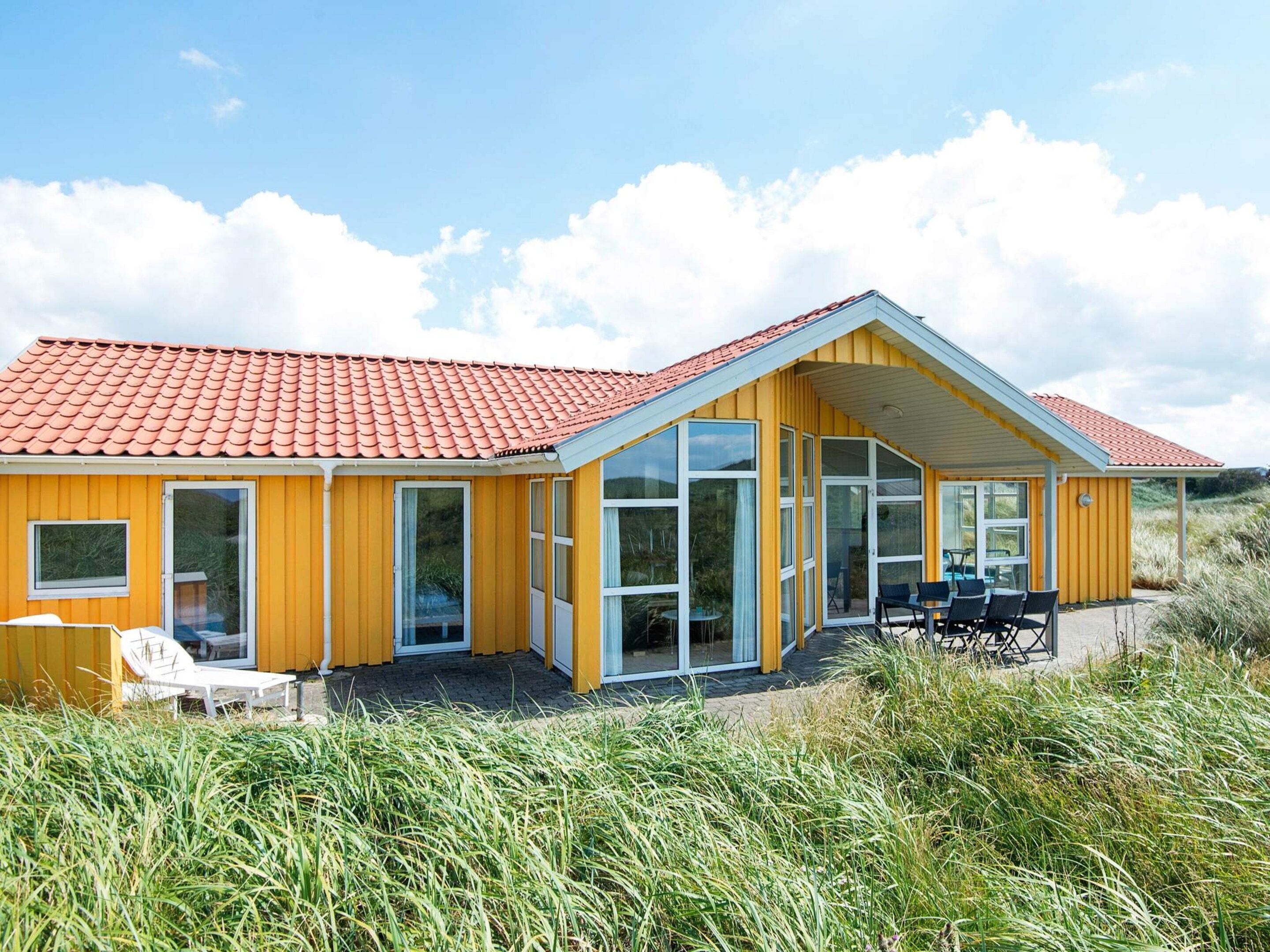 Ein gelbes Gebäude mit rotem Ziegeldach, großen Fenstern und einer Terrasse mit Gartenmöbeln.