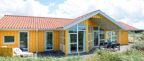 Ein gelbes Gebäude mit rotem Ziegeldach, großen Fenstern und einer Terrasse mit Gartenmöbeln.