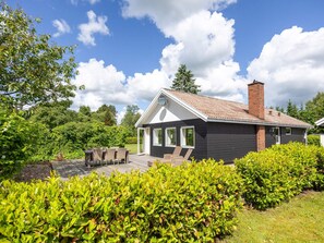 A modern house with a dark exterior, a tiled roof, and a chimney. It features a patio with outdoor furniture and a well-maintained garden.