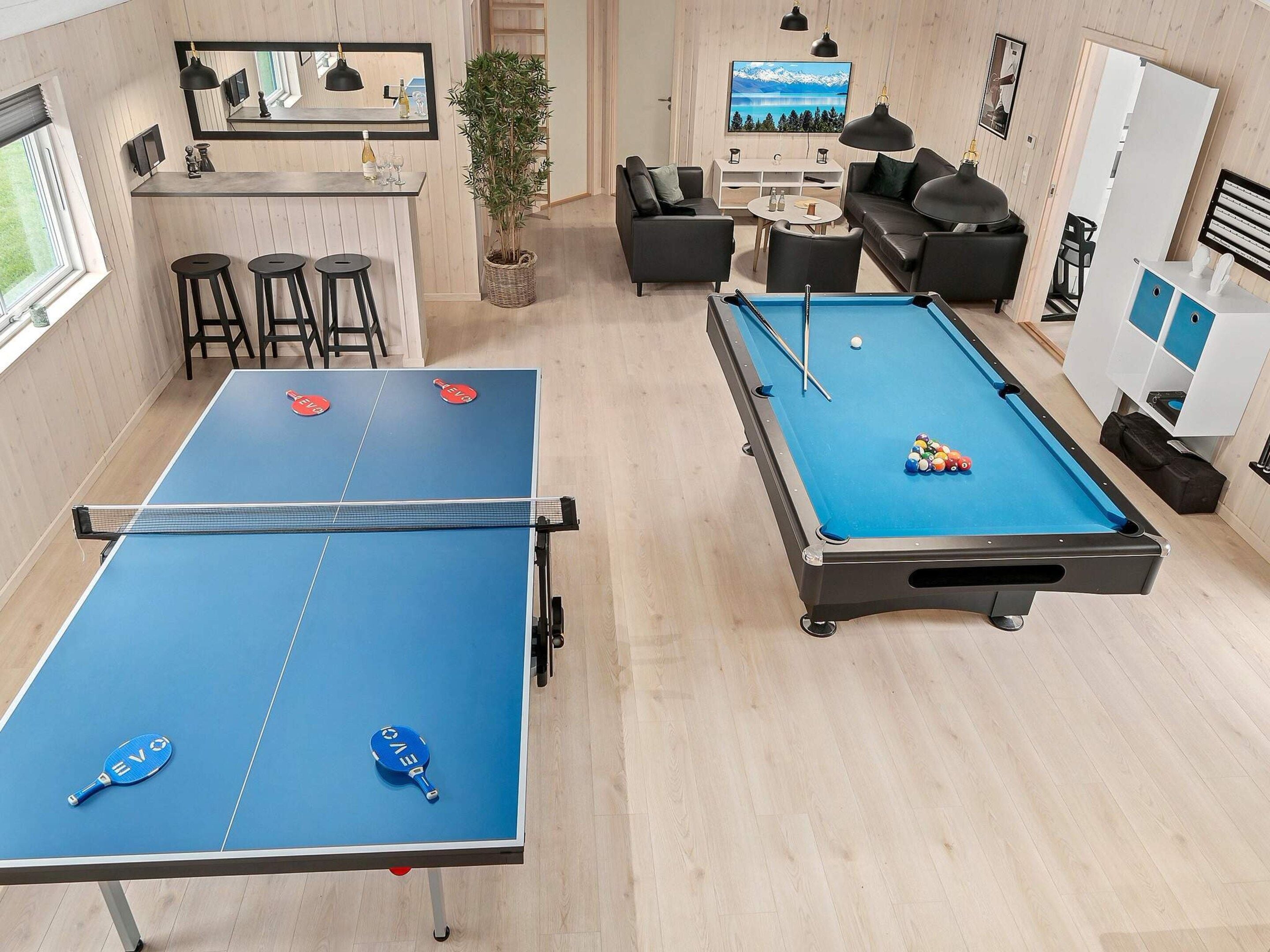A room with two pool tables, a bar area, a seating area with sofas, and a TV.
