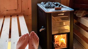 Un poêle à bois Harvia avec un feu qui crépite à l’intérieur, des pieds reposant devant, et une fenêtre en arrière-plan.