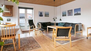 A spacious living room with wooden flooring, a black leather sofa, wooden chairs, a small table, and a large window with a view of a building outside. - 7 Person Holiday Home in Skagen (Skagen)