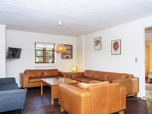 Ein Wohnzimmer mit einem Ecksofa, einem Couchtisch, einem an der Wand montierten Fernseher und einem Fenster mit Blick auf Bäume.