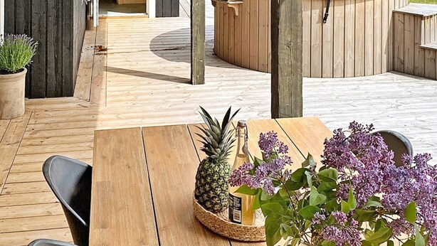 Une terrasse en bois avec un jacuzzi, une table ornée d’un ananas et de fleurs, et une plante en pot.