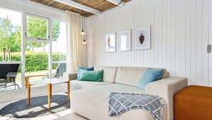 Un salon moderne avec un canapé, une table basse et un coin repas avec des chaises. De grandes fenêtres sont ornées de rideaux, le plafond est doté de poutres en bois et des tableaux encadrés décorent les murs.