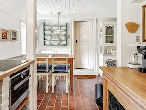 Uma cozinha com piso de azulejos, mesa de madeira, cadeiras, luminária pendente e uma porta que dá para outro cômodo.