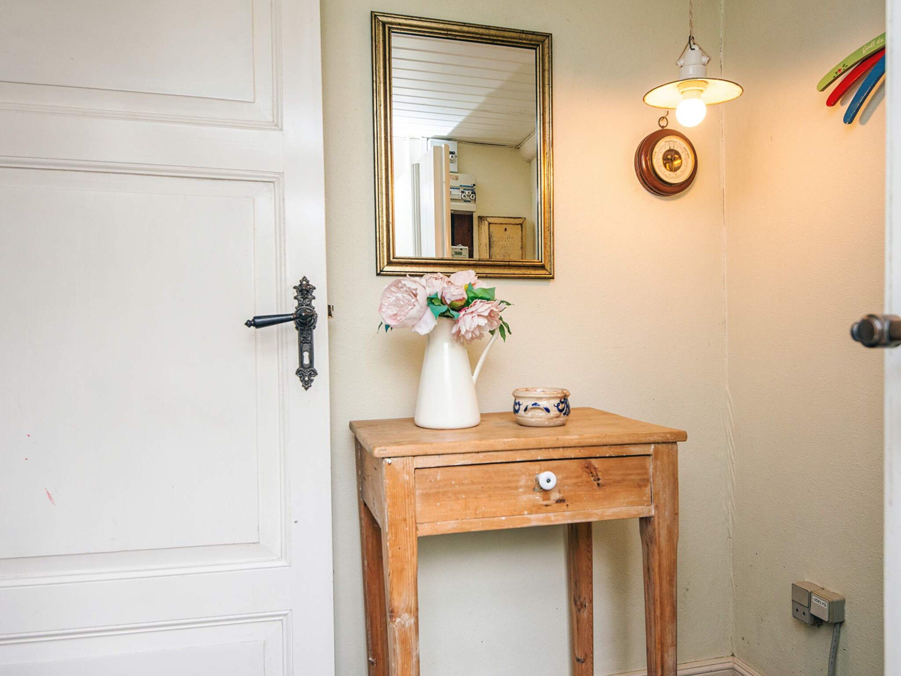 A white door with an ornate handle, a wooden bedside table with a white vase and a small ceramic container, a mirror with a gold frame, and a wall-mounted light fixture.