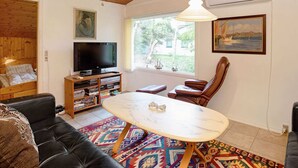 A cozy living room with a wooden ceiling, a television, a bookshelf, a leather sofa, a round table, and a large window with a view of greenery. - 6 Person Holiday Home in Dannemare (Dannemare)