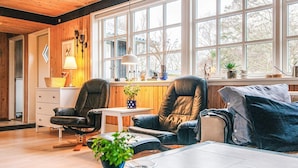 Ett mysigt vardagsrum med träväggar, ett stort fönster, en soffa, två fåtöljer, ett soffbord och en lampa.