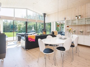 A modern living room with a fireplace, a dining area with white chairs, and a kitchen with glass cabinets. - Holiday Home in Glesborg (Glesborg)