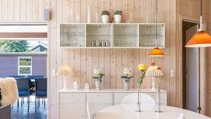 Eine moderne Küche mit weißen Schränken, ein Essbereich mit weißem Tisch und eine Holzwand mit Regalen, auf denen Glaswaren und Blumentöpfe präsentiert werden.