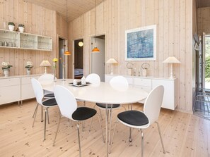 A modern dining area with a round white table, white chairs, and a wooden wall with a framed artwork. - Holiday Home in Glesborg (Glesborg)