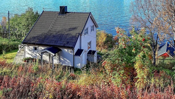 Una casa blanca con techo oscuro, rodeada de árboles y arbustos, con vista a un lago tranquilo y montañas al fondo.