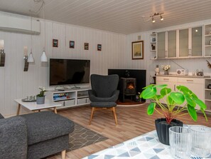 A modern living room with a television, a comfortable armchair, a sofa, a fireplace, and a kitchen area with cabinets and a radio. - 6 Person Holiday Home in Nordborg (Nordborg)